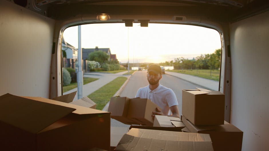 A man wearing a white polo shirt and glasses is seen standing outside the back of a van during a home relocation process, holding and loading cardboard boxes into the vehicle. The van's rear doors are open, revealing an interior filled with additional boxes and packing materials, such as cardboard containers and protective wrapping. The scene takes place in a suburban neighbourhood with houses, a sidewalk, and a lamp post visible along the street, illuminated by warm, late afternoon sunlight. The man from Man with Van Lambeth is engaged in a furniture transport or packing and moving activity, preparing items for transport as part of a professional removals service, with a focus on loading and logistical planning during the home relocation process.