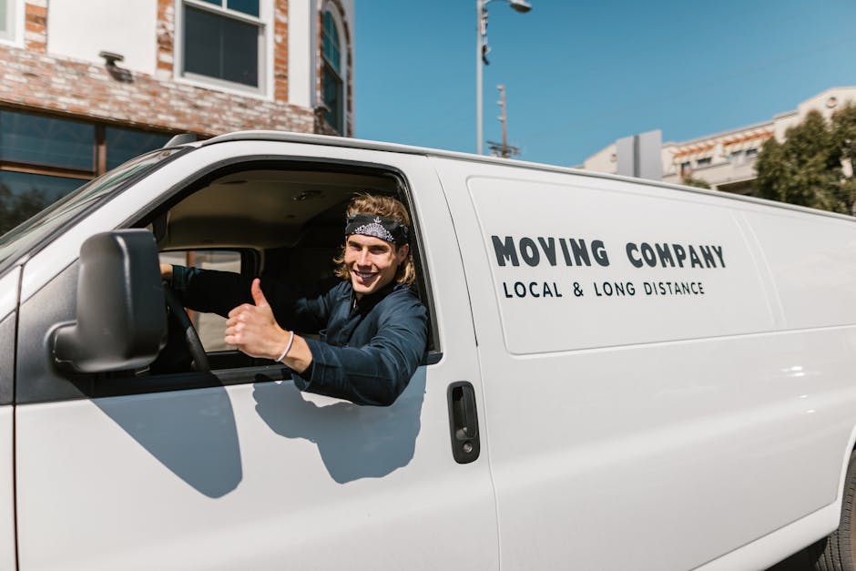 A young male mover with long hair, wearing a black bandana and dark blue work shirt, is sitting in the driver's seat of a white panel van labeled 'MOVING COMPANY LOCAL & LONG DISTANCE'. He is smiling and giving a thumbs-up gesture through the open window. The van is parked outdoors on a sunny day, with a modern building featuring large glass windows and a brick facade visible in the background. The scene captures the vehicle used for home relocation and furniture transport services provided by Man with Van Lambeth, indicating readiness for a house removal or moving job. The vehicle's side door is closed, and the environment suggests preparation for loading or unloading household items during a professional moving process.