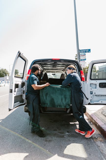 Two movers from Man with Van Lambeth are loading a large, dark green furniture piece into the open rear of a black van parked on a street in Brixton SW2. The furniture is covered with protective fabric or padding, and the movers are carefully maneuvering it on a trolley or dolly. The van's rear doors are wide open, revealing the spacious interior used for home relocation and furniture transport. In the background, there is a street sign reading 'Somerset' and a clear sky above, indicating good weather conditions for packing and moving services. The scene captures part of the pavement and roadside, highlighting the logistics involved in residential removals, especially for properties with narrow stairs, as referenced in the related guide.