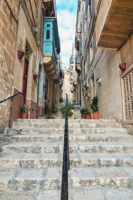 A young male mover with long hair, wearing a black bandana and dark blue work shirt, is sitting in the driver's seat of a white panel van labeled 'MOVING COMPANY LOCAL & LONG DISTANCE'. He is smiling and giving a thumbs-up gesture through the open window. The van is parked outdoors on a sunny day, with a modern building featuring large glass windows and a brick facade visible in the background. The scene captures the vehicle used for home relocation and furniture transport services provided by Man with Van Lambeth, indicating readiness for a house removal or moving job. The vehicle's side door is closed, and the environment suggests preparation for loading or unloading household items during a professional moving process.