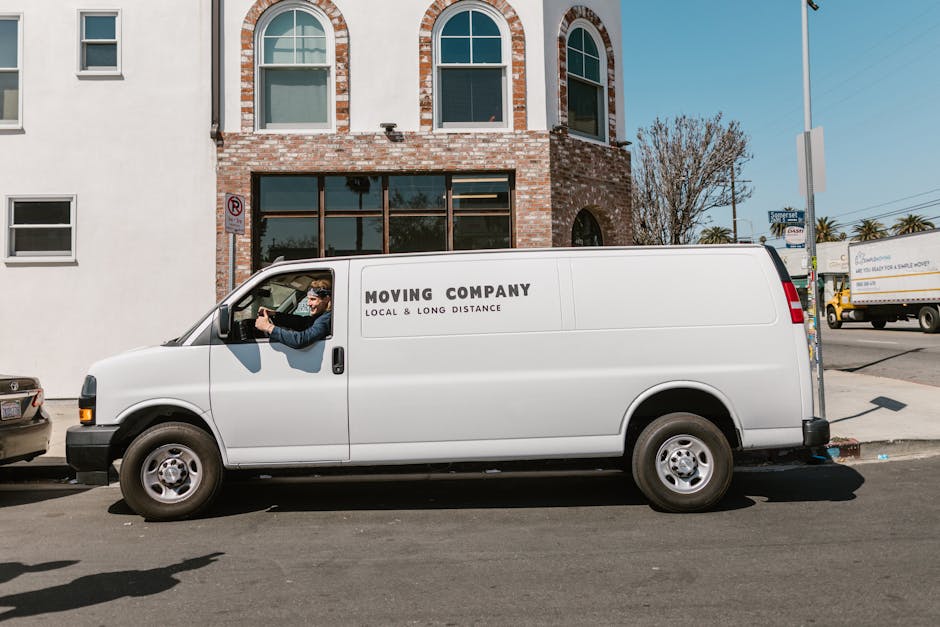 A white moving van with the text 'MOVING COMPANY LOCAL & LONG DISTANCE' printed on the side is parked parallel to a pavement in front of a brick building with three large arched windows. The driver, w