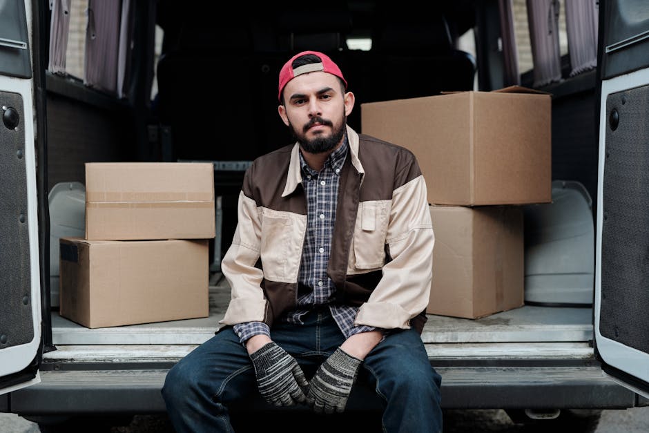 A professional mover from Man with Van Lambeth, wearing a white polo shirt and a black cap, stands outdoors beside a white moving van with its rear doors open. Inside the van, there are several stacke