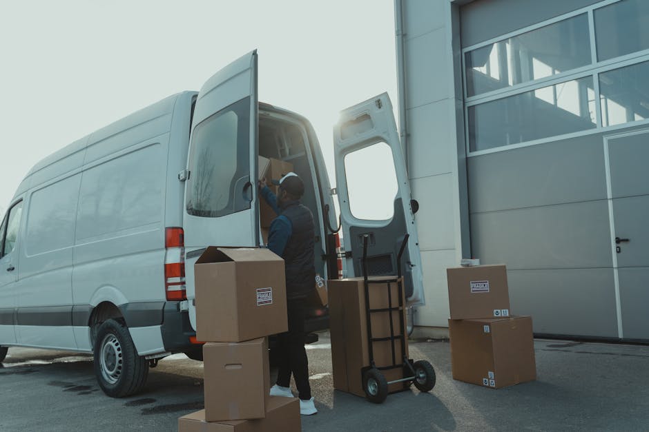 A worker loading cardboard boxes into the rear of a large white van parked outside a modern industrial building with large glass windows. The worker is wearing casual clothing and a cap, and is standing on the pavement, using a hand truck to assist with moving the boxes. Several additional boxes are placed on the ground nearby, some with labels indicating they are ready for transport. The van's rear doors are open, revealing a partially filled interior with more packed boxes and packing materials. Bright natural daylight illuminates the scene, with the sun positioned behind the van, creating a slight glare. This image illustrates the process of furniture transport, packing, and home relocation logistics handled by Man with Van Lambeth, as part of their removal services aligned with Lambeth Council's moving rules for large vans and suspensions.