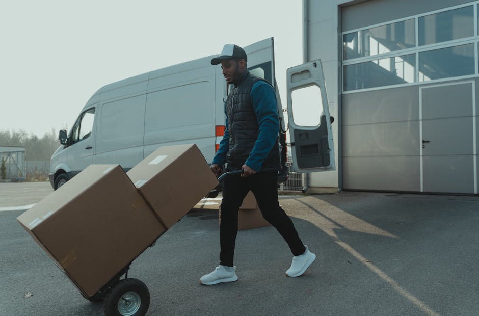 A man wearing a baseball cap, dark vest, blue long-sleeve shirt, black pants, and white sneakers is pushing a two-wheeled hand truck loaded with two large cardboard boxes outside a white cargo van. The van is parked near a modern, glass-fronted building with an open rear door, revealing some packaging materials and additional boxes inside. The scene takes place in a paved area with clear daylight, suggesting an organized home relocation or furniture transport process. Behind the van, there is a low fence and some trees in the distance. This image illustrates a typical loading and packing phase involved in house removals, supporting services like furniture transport and moving logistics, as provided by Man with Van Lambeth, aligned with local council moving rules for large vans and suspensions.