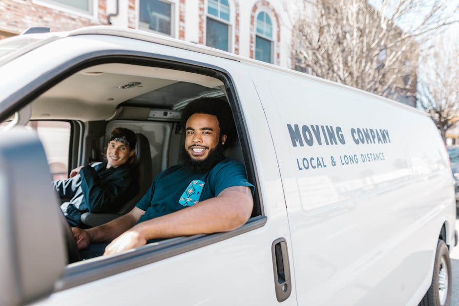 Inside a white moving van with its side door open, two movers from Man with Van Lambeth are visible. The driver, a man with dark curly hair and a beard, is sitting in the front seat, smiling at the camera. The passenger, a woman with dark hair pulled back and wearing a dark jacket and headband, is seated behind him, also smiling. The van features the words 'MOVING COMPANY' and 'LOCAL & LONG DISTANCE' written on the side. The interior of the van shows some packing supplies, such as cardboard boxes and plastic wrapping, suggesting they are in the process of loading or unloading items for a home relocation. The setting appears to be a residential area with trees and multi-storey buildings with large windows visible outside. The lighting indicates daytime, and the overall scene captures the preparation for furniture transport and packing in a professional move, aligning with the regulations regarding permits for removals in Lambeth Council zones.