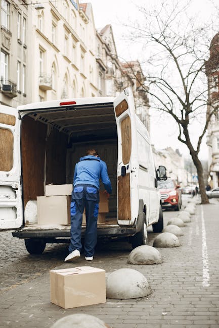 A narrow residential street in Lambeth lined with terraced houses featuring brick and white-painted facades, with several potted plants and small trees positioned along the pavement. In the center of the scene, a white van with a spare tire mounted on the rear is parked, indicating a home relocation or moving activity. The street appears to be in daytime with natural light illuminating the scene, and some buildings under construction or renovation are visible on the left side, covered with scaffolding and protective sheeting. The arrangement of objects and vehicles suggests a typical environment where house removals and furniture transport may involve navigating narrow streets with potential access restrictions, reflective of challenges related to permits and street access in Lambeth, as addressed by Man with Van Lambeth's removals services.
