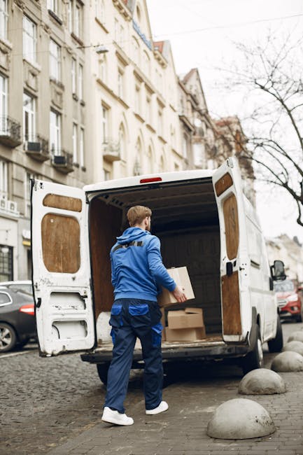 A man wearing a blue jacket and dark trousers is standing at the back of a white moving van parked on a cobblestone street in Lambeth. The van's rear doors are open, revealing the interior lined with wooden panels and filled with several packed cardboard boxes in various sizes, some wrapped in plastic or fabric covers. The man is holding a cardboard box, preparing to load it into the van, indicating the process of home relocation or furniture transport. The street is lined with multi-storey residential buildings with ornate facades, and parked cars are visible along the pavement. The scene is set during daytime, with overcast skies and leafless trees in the background, suggesting a winter season. This image illustrates the logistics involved in packing and loading for house removals, with a focus on professional moving services like those provided by Man with Van Lambeth, supporting same-day and last-minute move solutions.