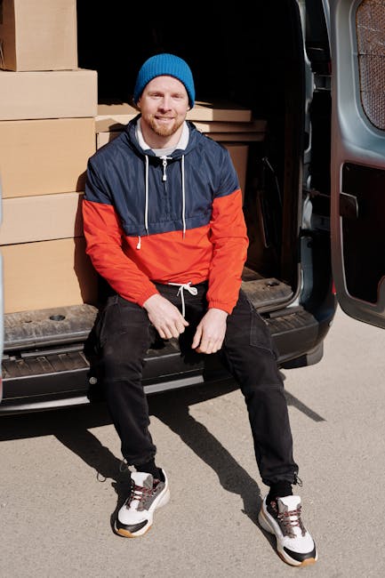 A man with a friendly expression, wearing a blue beanie, navy and red jacket, black pants, and sneakers, is sitting on the open rear door of a black van. The van's interior is visible behind him, showing stacked cardboard boxes of various sizes, some wrapped in plastic or fabric padding for protection during transportation. The man is positioned on a paved surface, with the side door of the van open, revealing the loading area where boxes are being unloaded or prepared for home relocation. The natural lighting indicates daytime, and the setting reflects a professional furniture transport or moving process associated with house removals or estate moves. The image is connected to Man with Van Lambeth's services, emphasizing packing, loading, and vehicle loading stages in a typical house moving scenario.