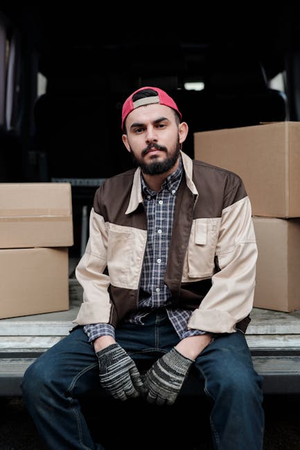 A young man with a beard wearing a red cap, a checked shirt, and a beige and brown jacket is sitting on the edge of a vehicle's open trunk during a home relocation process. Behind him, there are several plain cardboard boxes stacked inside the vehicle, which appears to be a van or a moving truck, with additional packing materials and possibly some foam or fabric protective coverings partially visible. The man is wearing grey and black striped gloves, and he looks directly at the camera with a neutral expression. The scene suggests he is involved in furniture transport and loading activities typical of house removals, with the vehicle positioned outdoors on a driveway or street, under natural lighting. Man with Van Lambeth specializes in removals, and this image illustrates the loading process of packing and moving items for a house move, aligning with the context of the services offered in the Vauxhall SW8 area.