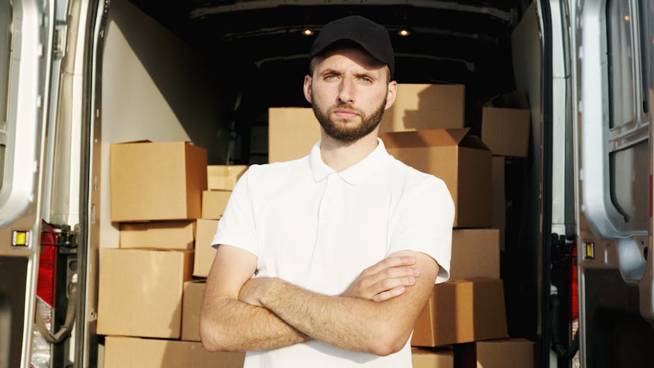A man with a friendly expression, wearing a blue beanie, navy and red jacket, black pants, and sneakers, is sitting on the open rear door of a black van. The van's interior is visible behind him, showing stacked cardboard boxes of various sizes, some wrapped in plastic or fabric padding for protection during transportation. The man is positioned on a paved surface, with the side door of the van open, revealing the loading area where boxes are being unloaded or prepared for home relocation. The natural lighting indicates daytime, and the setting reflects a professional furniture transport or moving process associated with house removals or estate moves. The image is connected to Man with Van Lambeth's services, emphasizing packing, loading, and vehicle loading stages in a typical house moving scenario.