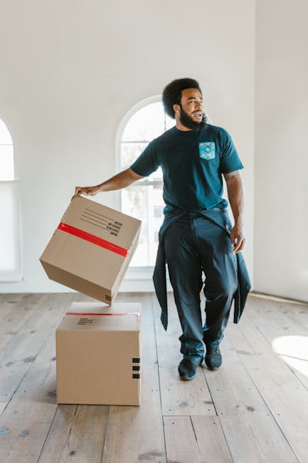 A man with dark curly hair and a beard is inside a bright, empty room with white walls and large arched windows, which let in natural light. He is dressed in a blue T-shirt and dark trousers, and stands next to a stack of two cardboard moving boxes. One box is placed on the floor while the other is being held in his right hand, slightly lifted and angled for placement or carrying. Both boxes are sealed with packing tape, with the top box featuring red tape around its middle. The room has a light wooden floor, and the scene appears to depict a home relocation or furniture transport process, with the man preparing to move or load the boxes. This visual aligns with the services of [COMPANY_NAME] and supports the context of house removals, packing, and moving logistics as discussed in the Waterloo station removals access tips for Lambeth moves page of manwithvanlambeth.org.