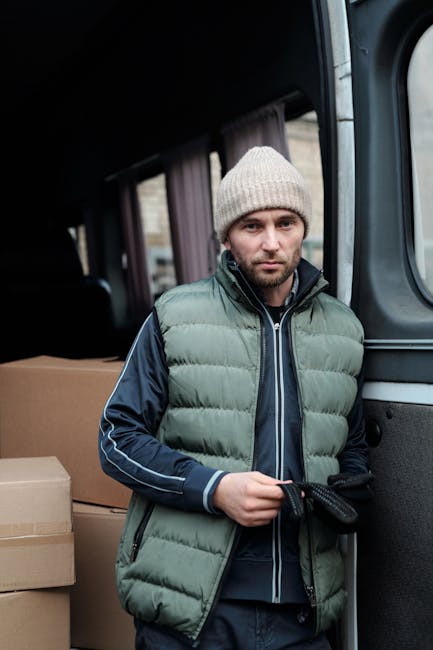 A man dressed in a light grey knitted beanie, green padded vest, and navy blue tracksuit top, standing inside a van with the sliding door open. Around him, there are several cardboard boxes, some sealed with packing tape, indicating an ongoing home relocation or furniture transport process. The background shows the interior of the van with dark upholstery and window curtains, suggesting a professional removals setting. The man is holding a piece of rope or strap, likely used for securing items during packing or loading. This scene reflects the logistics involved in furniture transport and packing during house removals, highlighting the detailed process of loading and securing belongings before moving, as part of a comprehensive moving service provided by Man with Van Lambeth.